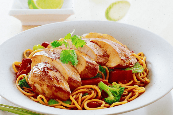 Teriyaki chicken and noodle stir fry in a white bowl with lime on the side.