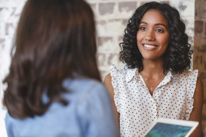 Two business women having a conversation. One person has her back to us. She is holding a digital tablet.