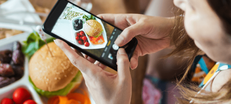 A woman takes a photo of a healthy homemade burger and bright salad on her mobile phone, the shot peers down from over her left shoulder