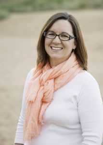 Woman with long brown hair and glasses wearing a pink scarf and white top smiling at the camera