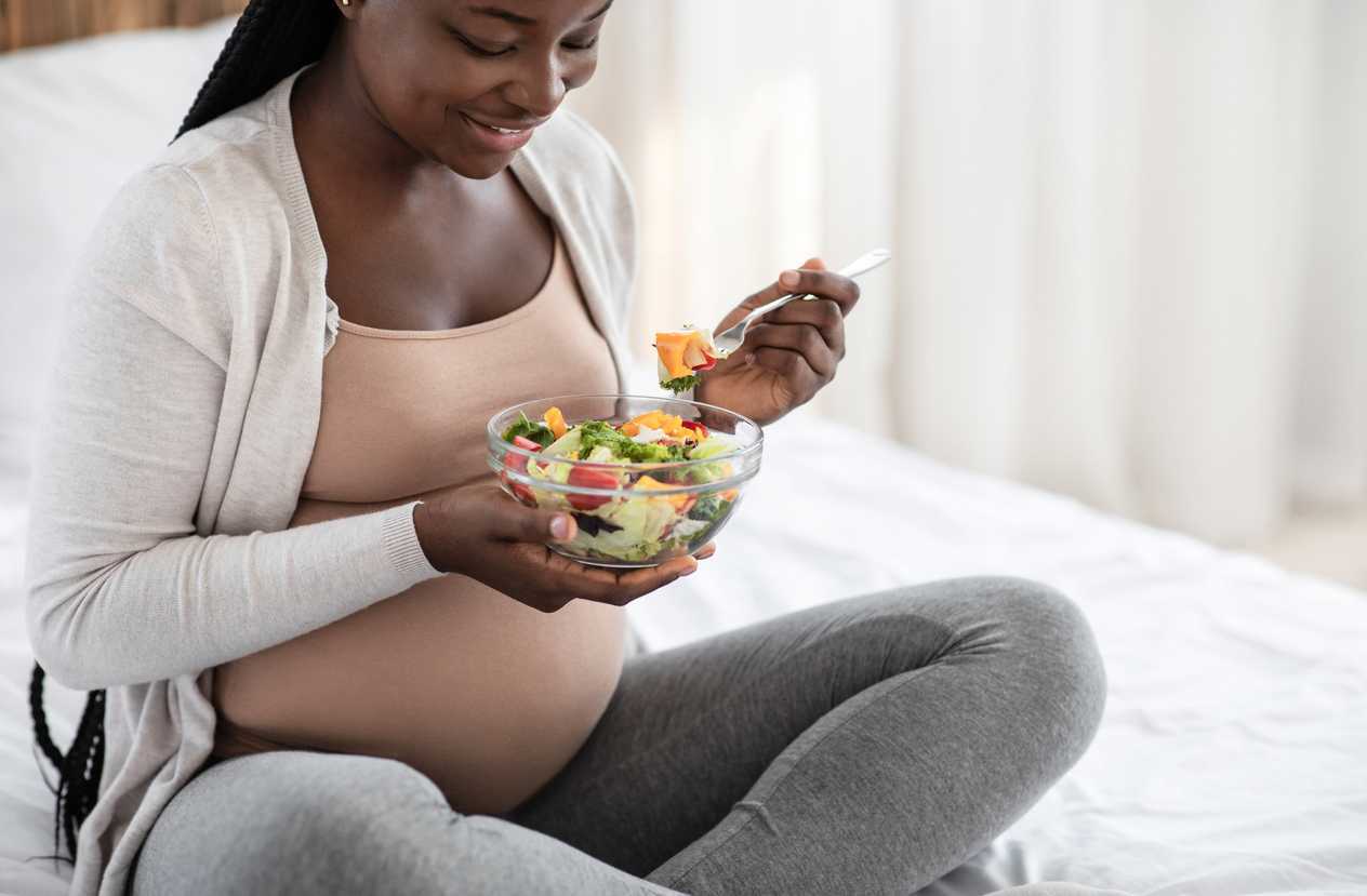 Pregnant woman sitting on bed eating salad
