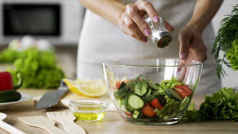 Woman adding salt in vegetable salad glass bowl