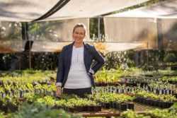 Dr Kristen MacKenzie-Shalders standing in a greenhouse among potted plants, wearing a white shirt and dark jacket, with sunlight filtering through shade cloths.