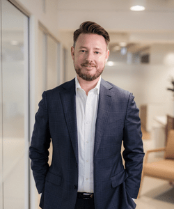 White man standing in office wearing grey suite with white shirt. Hands in pockets looking happy.
